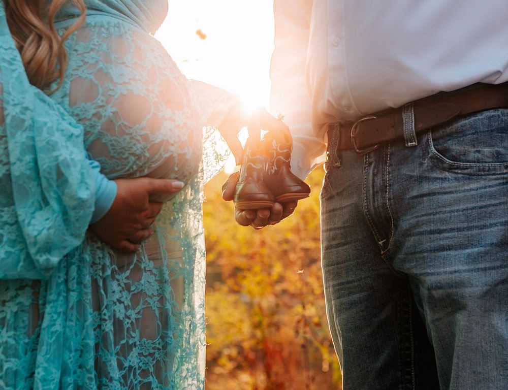 Pregnant couple holding hands in golden autumn sunlight, woman in teal lace maternity dress, man in jeans with leather belt.