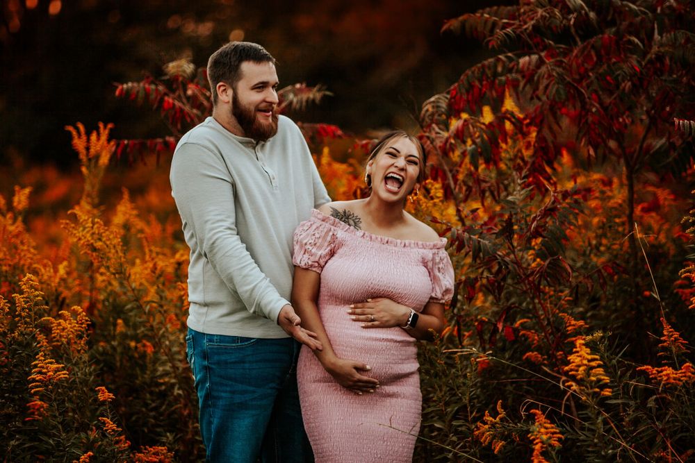 Couple laughing together in autumn foliage, pregnant woman in pink dress cradling baby bump during fall maternity photo session.