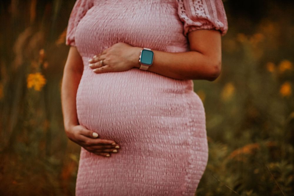 Pregnant woman in pink dress cradling baby bump outdoors in a field of wildflowers, wearing a teal smartwatch.