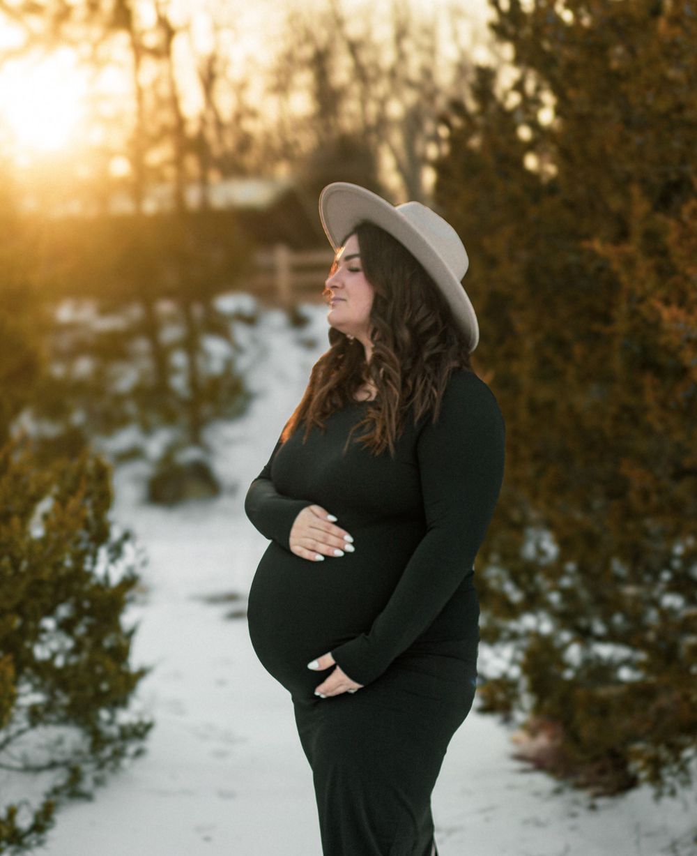 Pregnant woman in black dress and tan hat poses outdoors in snow at golden hour sunset.