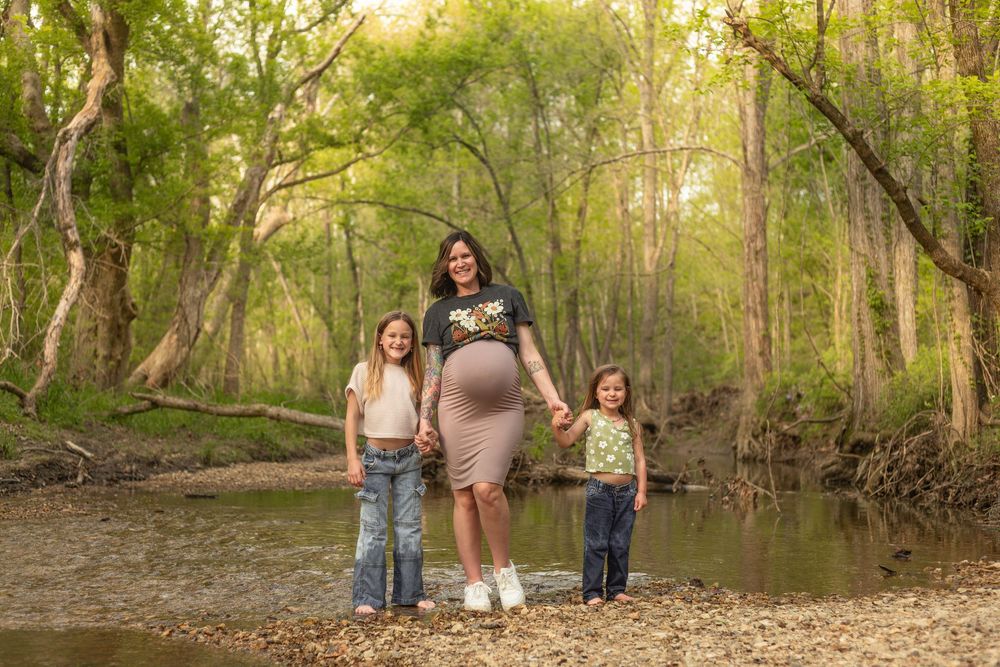 Pregnant woman stands outdoors by a creek with two young daughters in a lush green forest setting.
