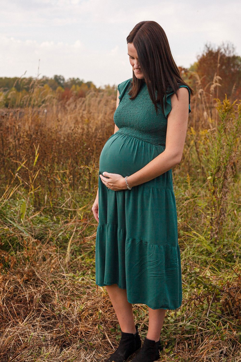 Pregnant woman in green dress cradling baby bump in autumn field.