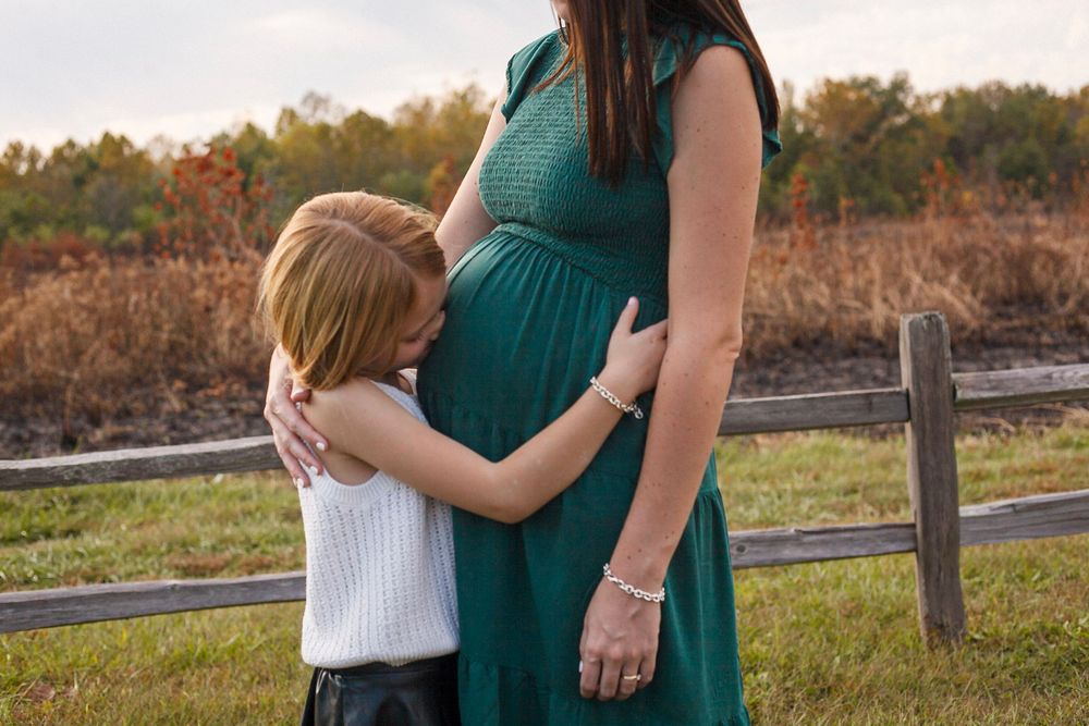 Pregnant woman in teal dress outdoors with child hugging her baby bump near a wooden fence.