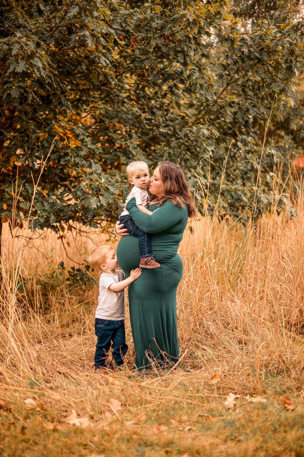 Pregnant woman in green dress holds toddler while another child hugs her belly in an autumn field.