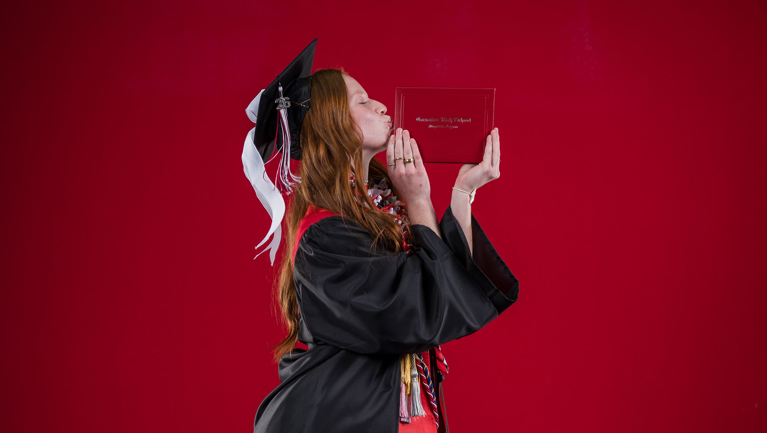 Female graduate in black cap and gown kisses diploma against vibrant red background.