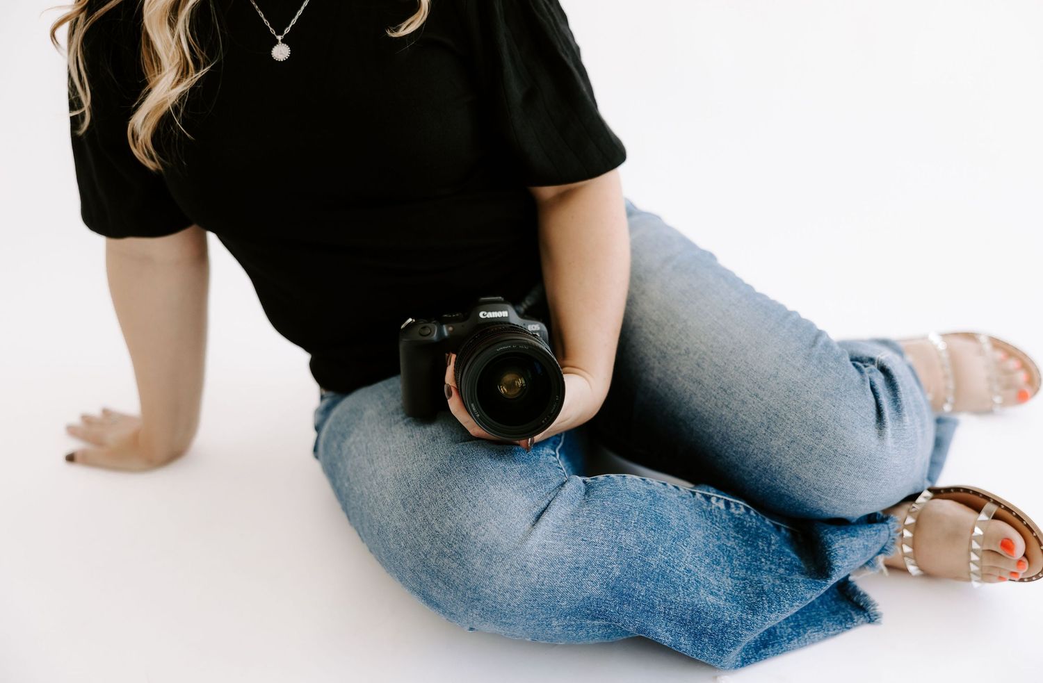 Woman in black top and jeans sitting on a white floor holding a professional DSLR camera.