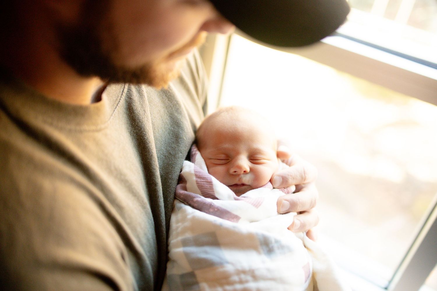 A bearded father in a gray shirt tenderly holds his sleeping newborn baby wrapped in a white blanket near a window.