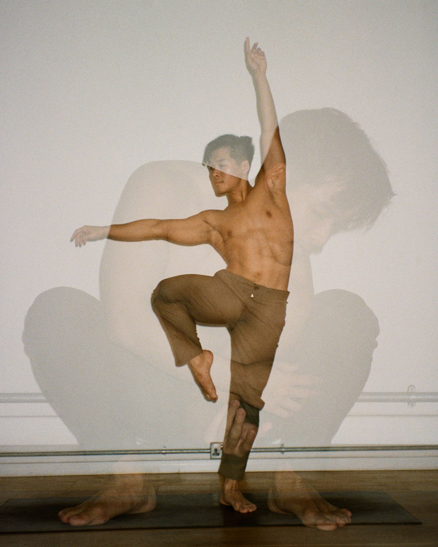 Shirtless male dancer balancing on one leg with arm raised in an artistic double exposure yoga pose.
