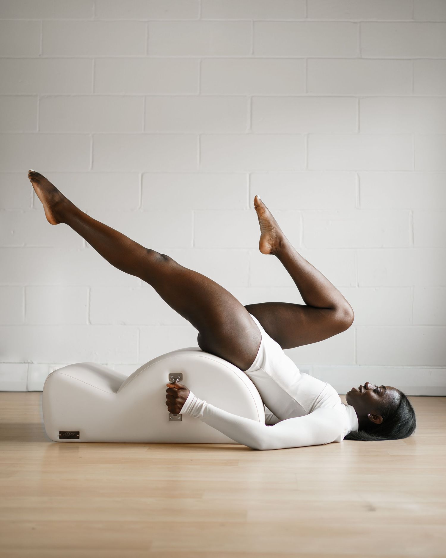 A person lying on a white curved Pilates arc prop, performing a leg raise exercise on a wooden floor.