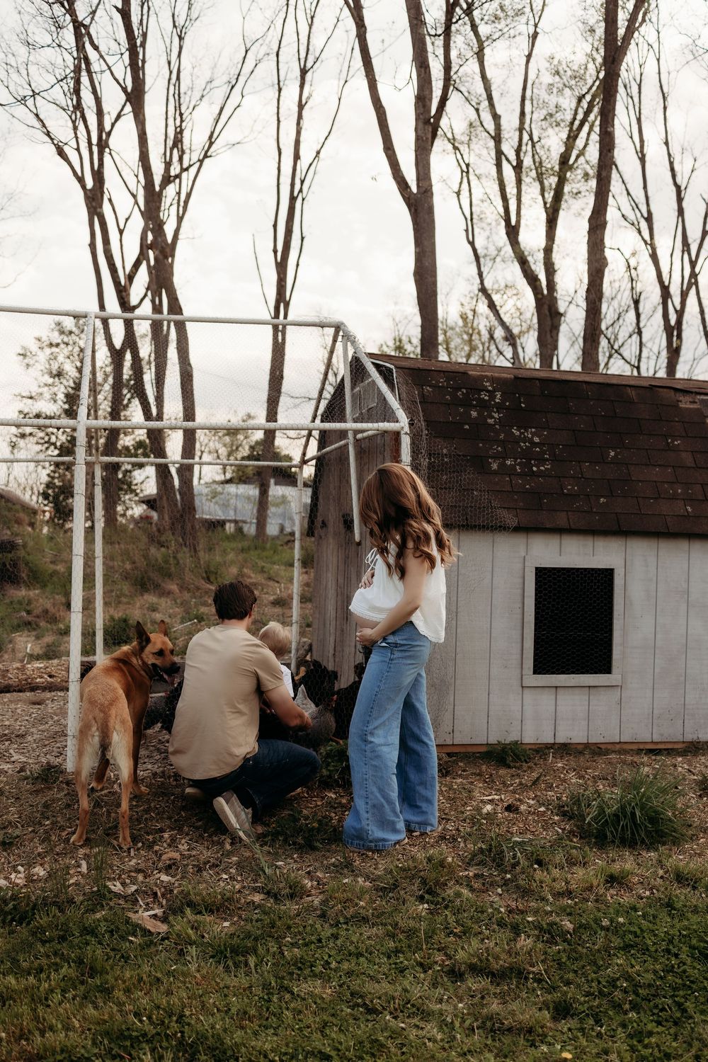 Family with dog exploring a rustic chicken coop in a wooded backyard during autumn.