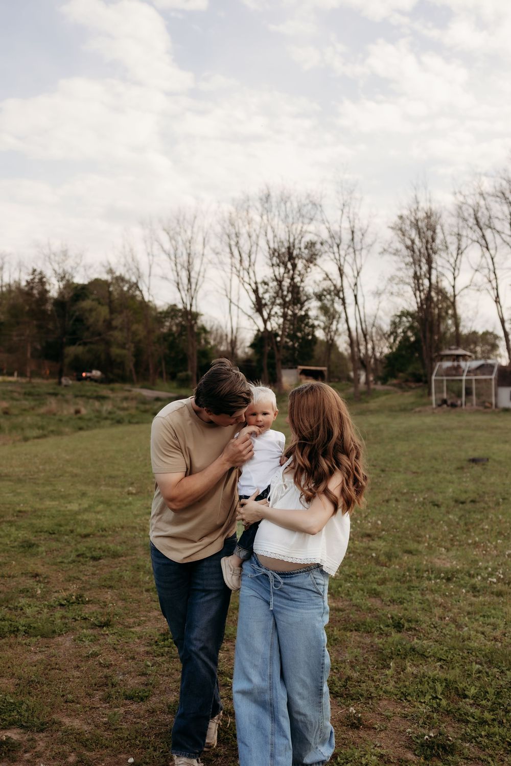 Young family with baby enjoying outdoor photo session in green field with trees in background.