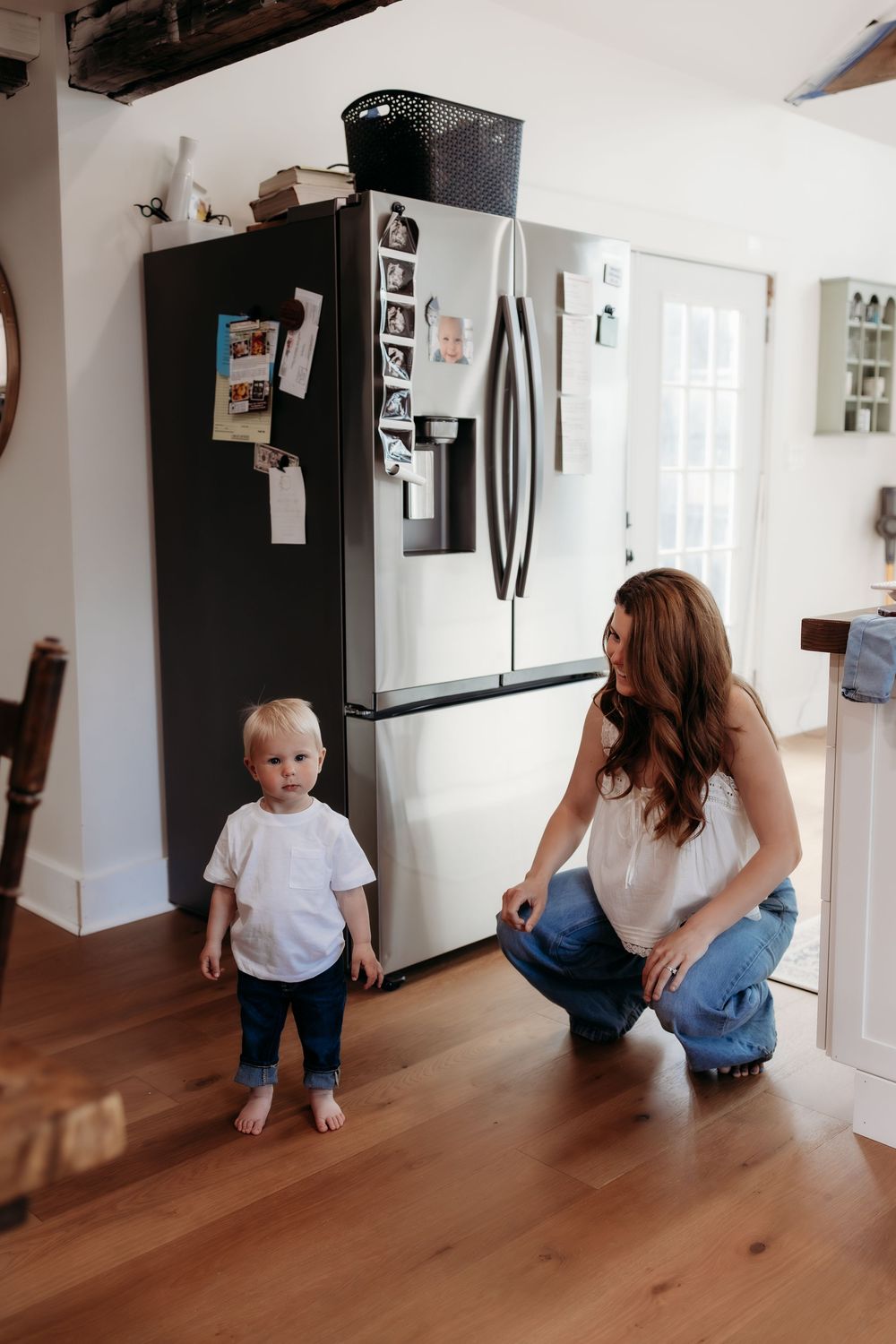 Mother kneels smiling at barefoot toddler standing in modern kitchen.