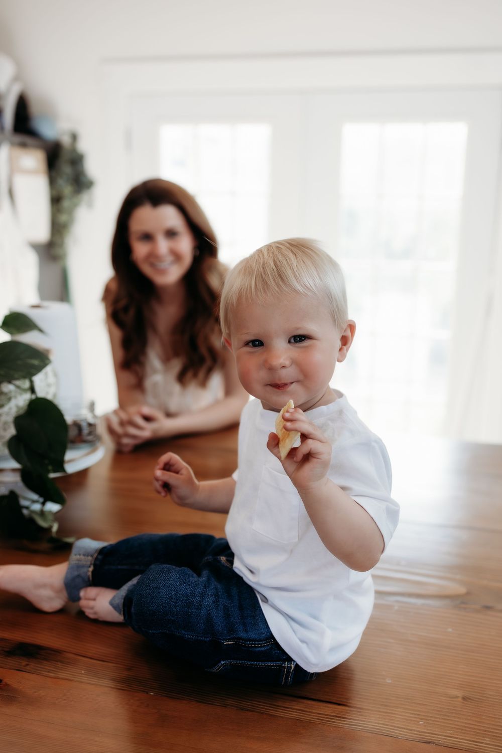 Toddler in white shirt sitting on hardwood floor eating a snack while mother smiles in background.