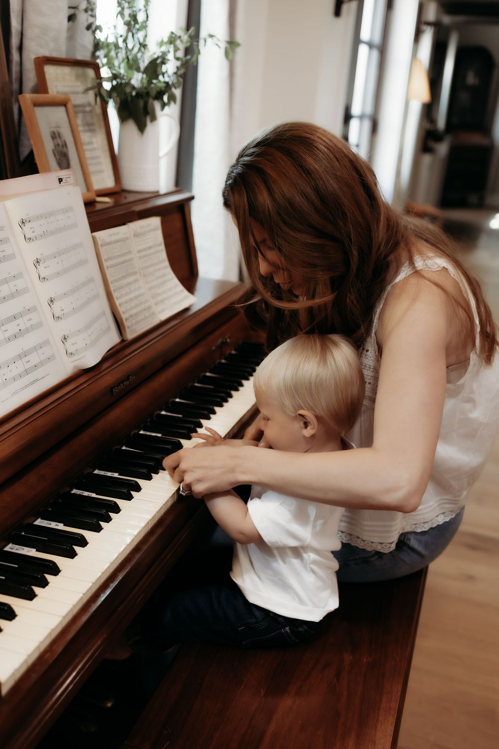 A mother helps her young toddler play piano keys while sheet music rests on a wooden upright piano.