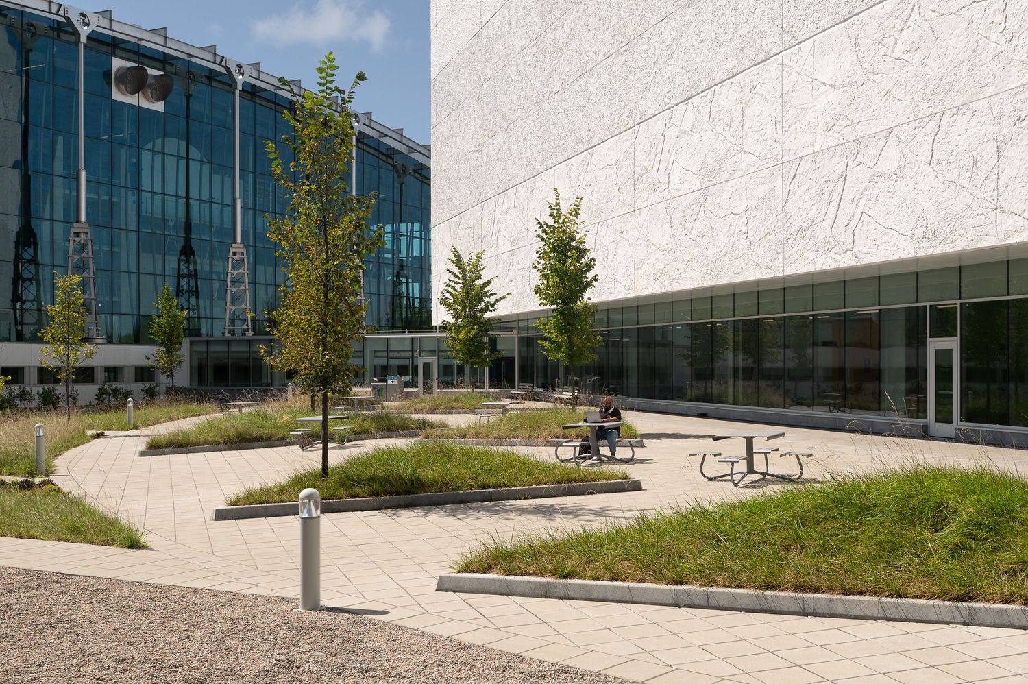 Modern office campus courtyard with glass buildings, landscaped greenery, curved pathways, and outdoor seating area.
