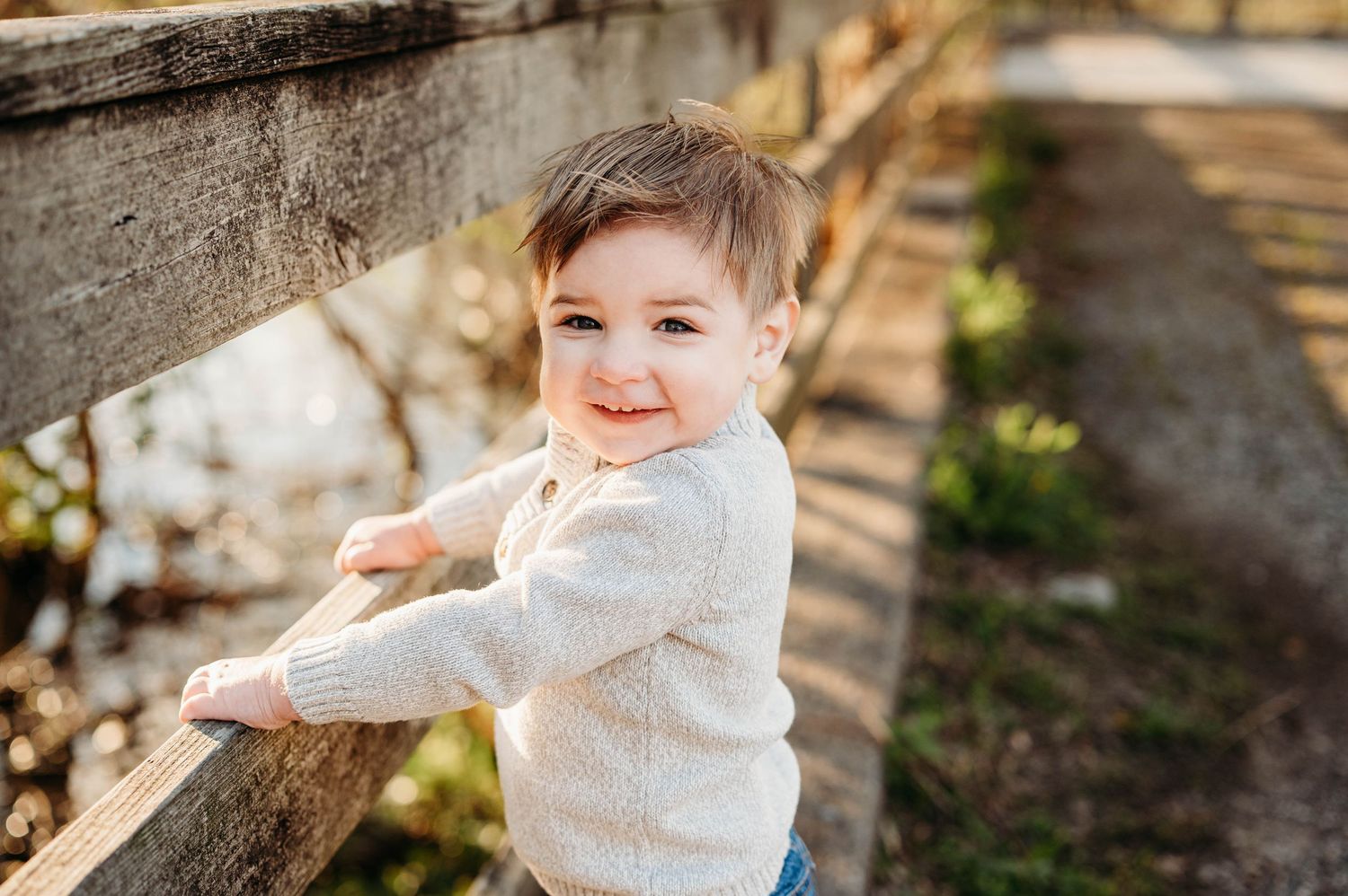 A happy toddler boy runs toward the camera in a grassy yard while his smiling parents stand together in the background.