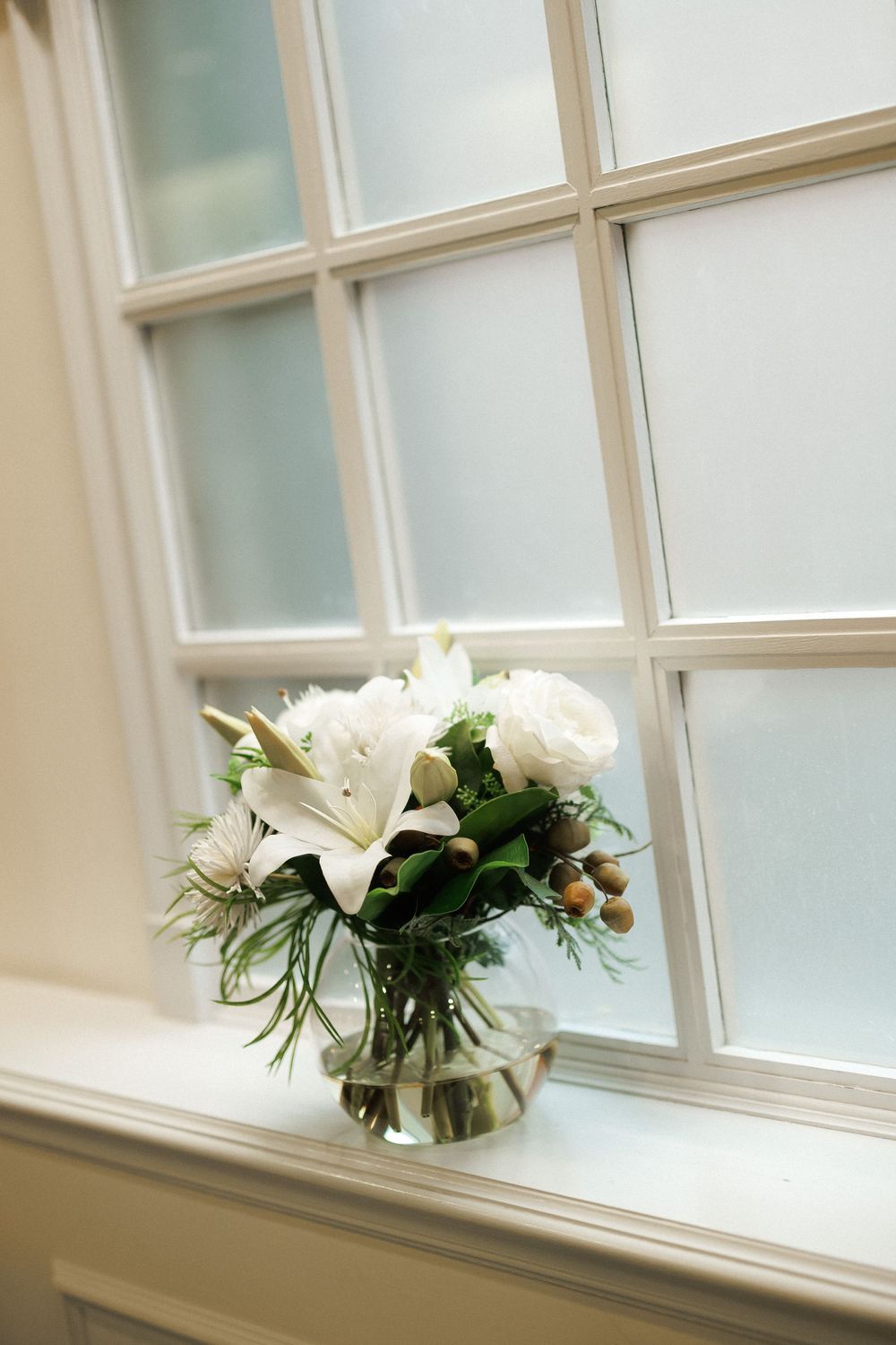 White lily and rose bouquet in glass vase on windowsill with frosted glass window backdrop.