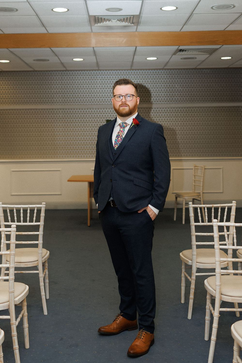 A bearded man in a navy suit with floral tie and brown shoes stands between white chairs in an elegant event room.