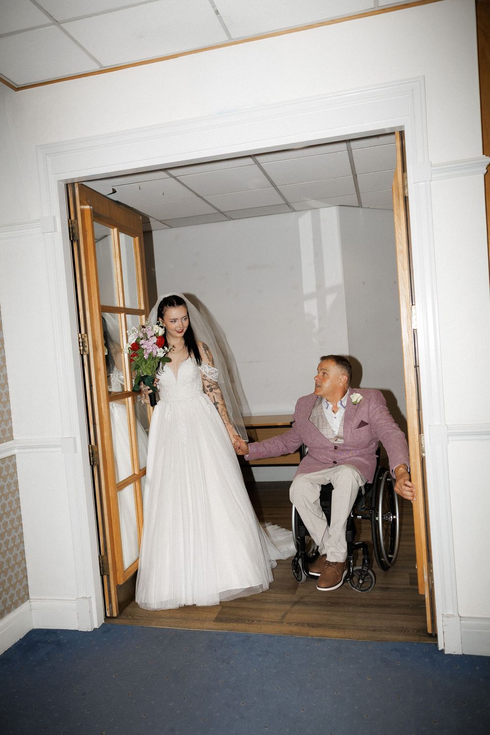 Bride in white gown holds bouquet while groom in pink suit sits in wheelchair, holding hands at wedding venue doorway.