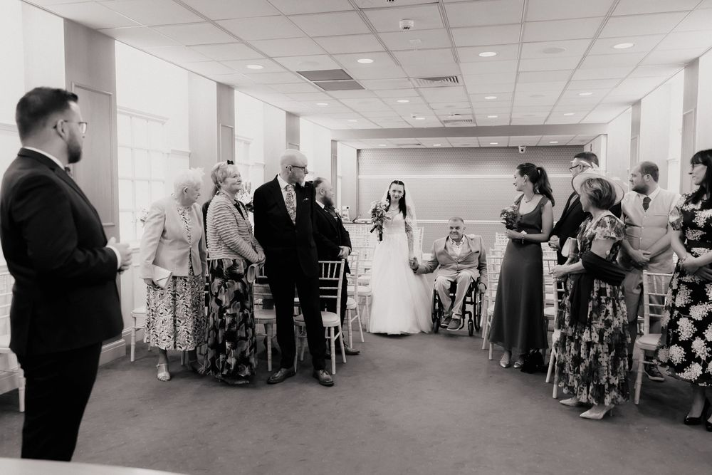 Black and white wedding ceremony photo showing bride walking down the aisle with guests standing in an elegant indoor venue.