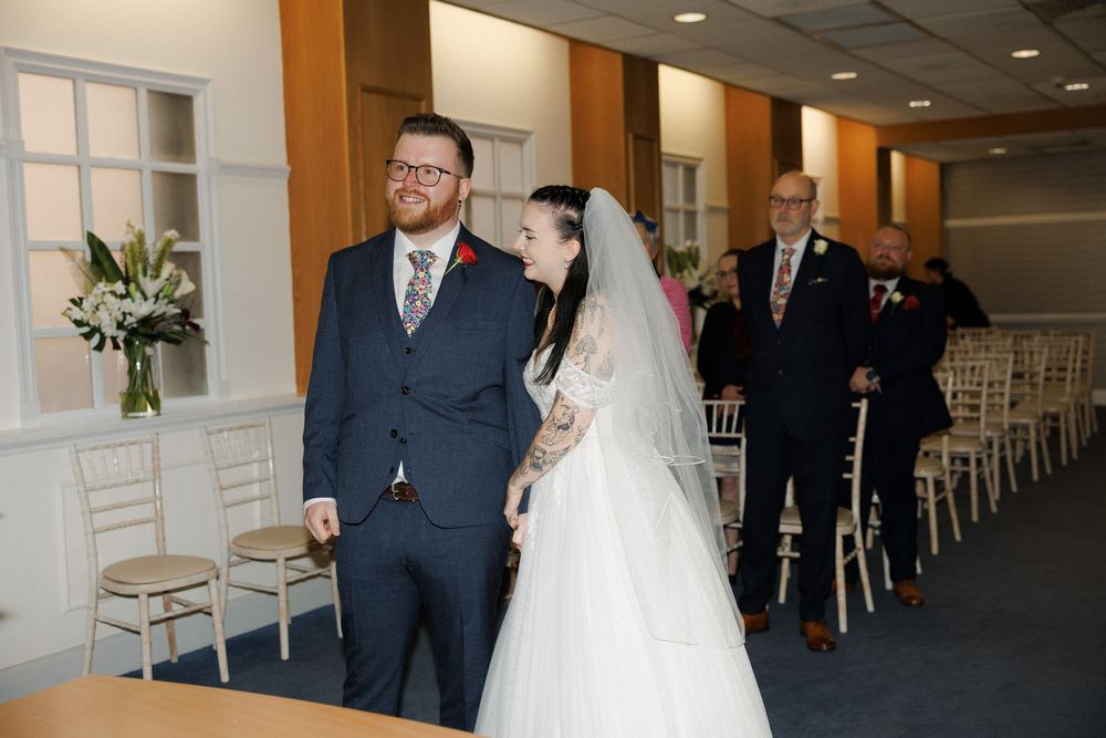 Bride in white lace gown and veil walks down aisle with groom in navy suit at elegant wedding ceremony.