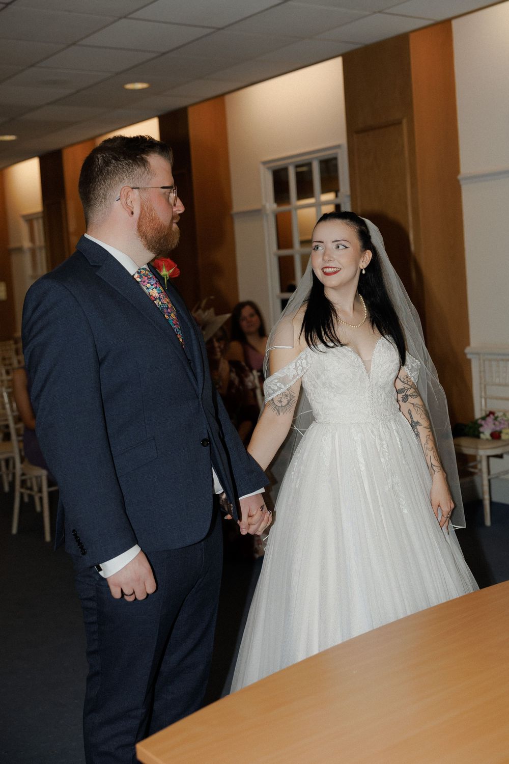 A bride in a white gown and veil holds hands with a groom in a navy suit at their wedding ceremony.