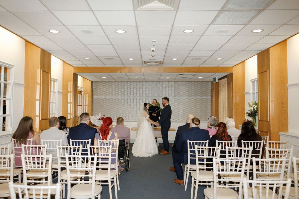 Elegant indoor wedding ceremony with guests seated on white chairs facing the couple at the altar in a bright venue.