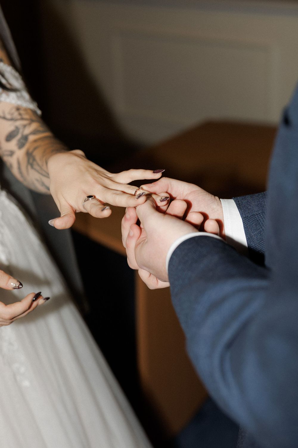 Groom placing a wedding ring on bride's finger during ceremony, bride has tattoo and black nail polish.