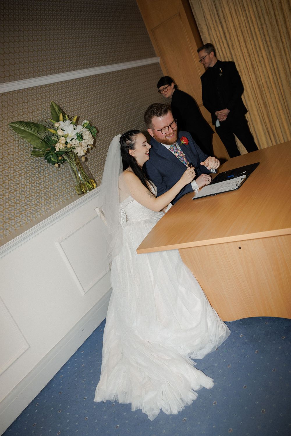 Bride and groom laughing together while signing wedding register at a wooden desk during their ceremony.