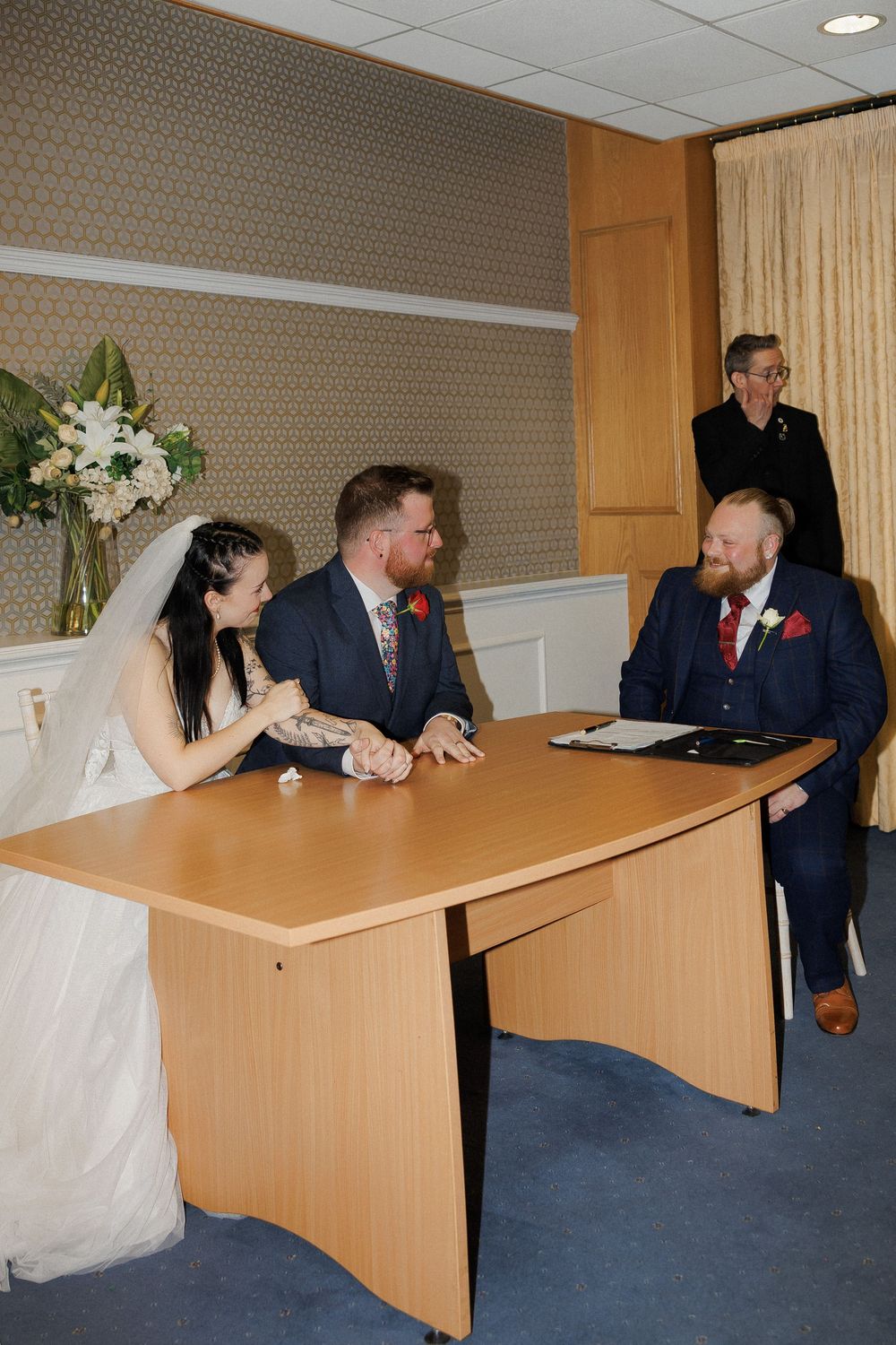 Bride and groom signing wedding documents at a table with a witness in a registry office ceremony room.