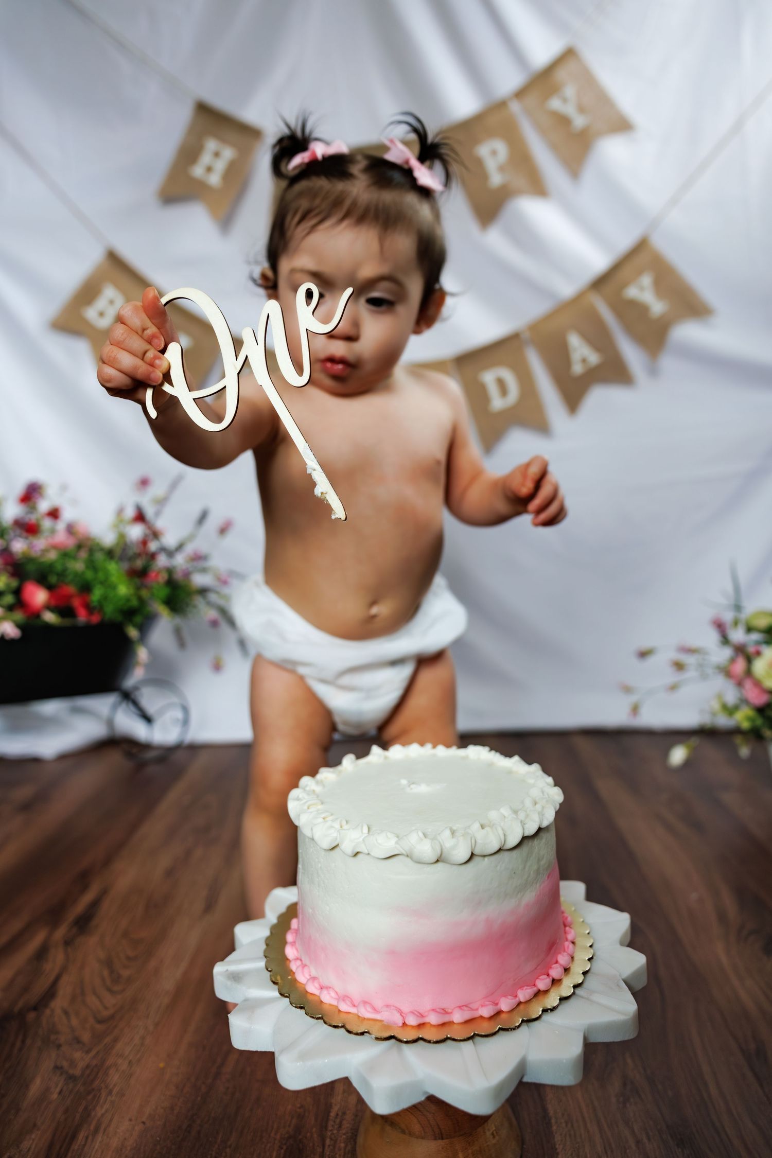 Baby in diaper holds "One" cake topper at first birthday party with pink ombre smash cake.