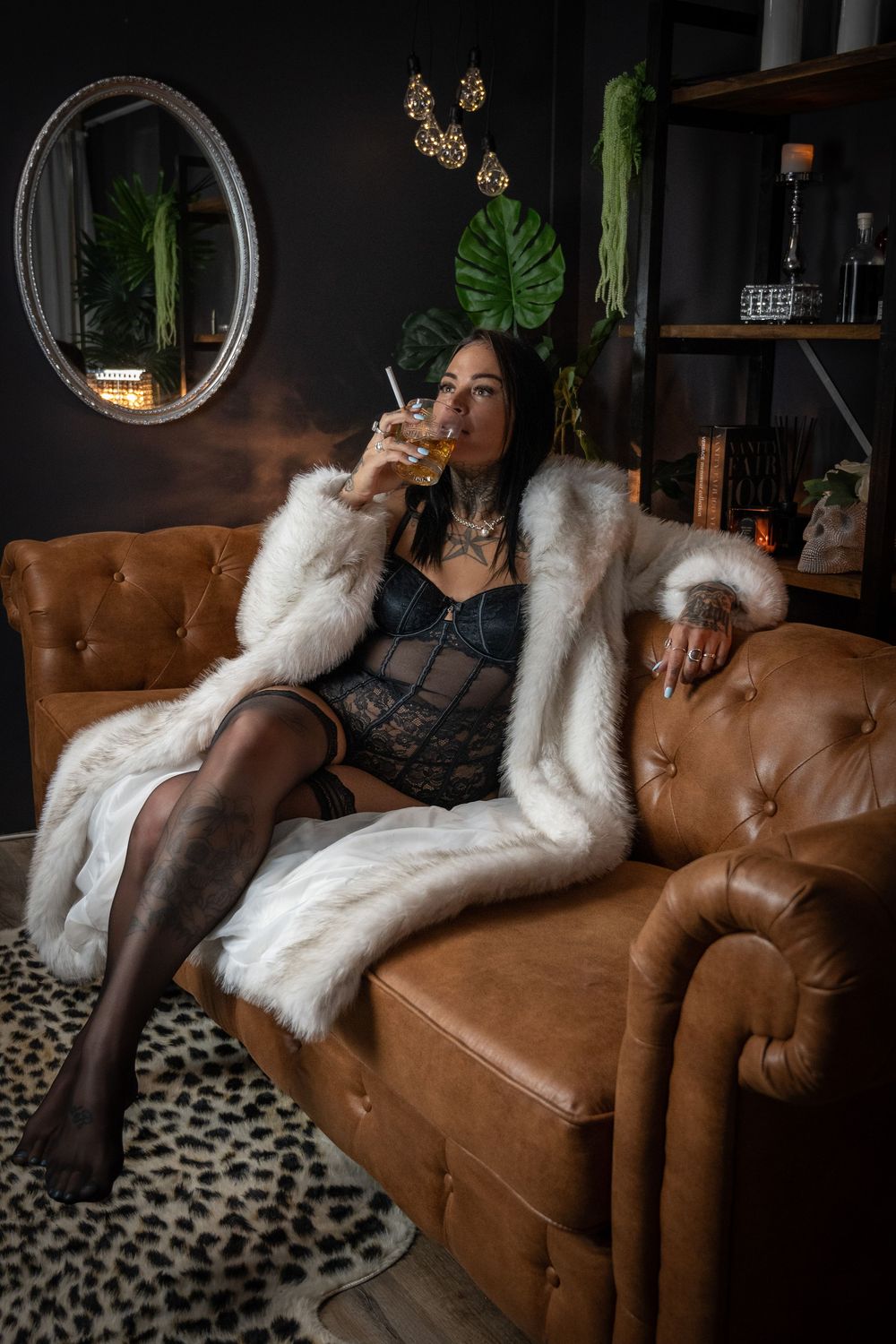 Woman in white fur coat and black lingerie sipping a cocktail on a brown leather tufted sofa in a dark luxe interior.