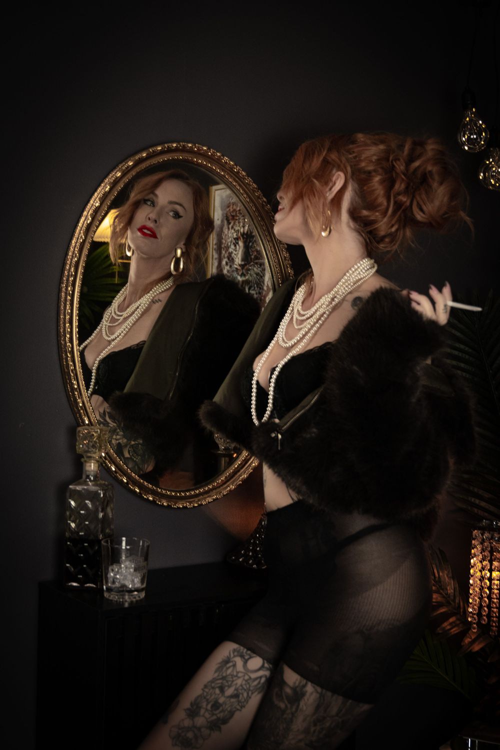 Glamorous redhead woman in black lingerie and pearls poses beside ornate gold mirror in vintage noir style setting.