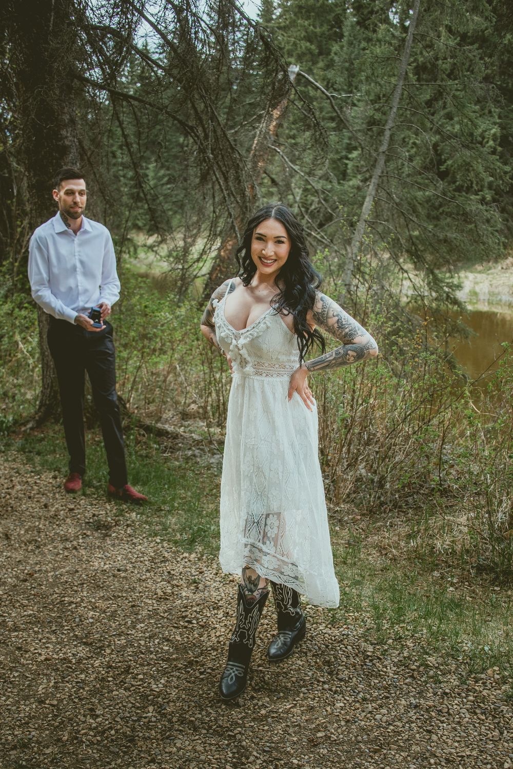Bride in white lace dress and cowboy boots smiles outdoors as groom stands behind her in forest setting.