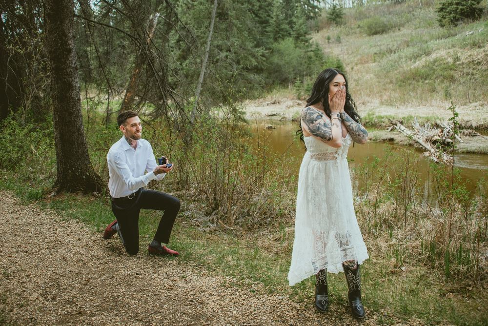 Man proposes on one knee to surprised woman in white dress outdoors near a creek in a lush green forest setting.