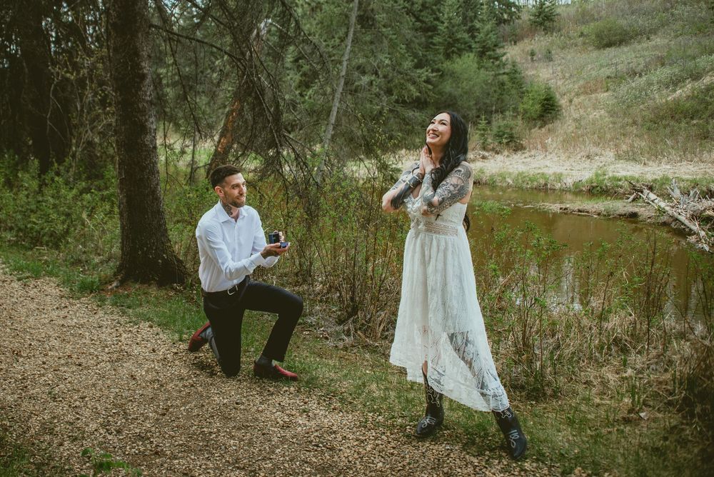 Man kneeling on one knee proposing to woman in white lace dress near forest creek outdoors.