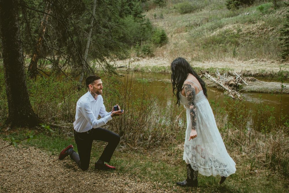 Romantic outdoor marriage proposal with man on one knee showing ring box to woman in white dress.