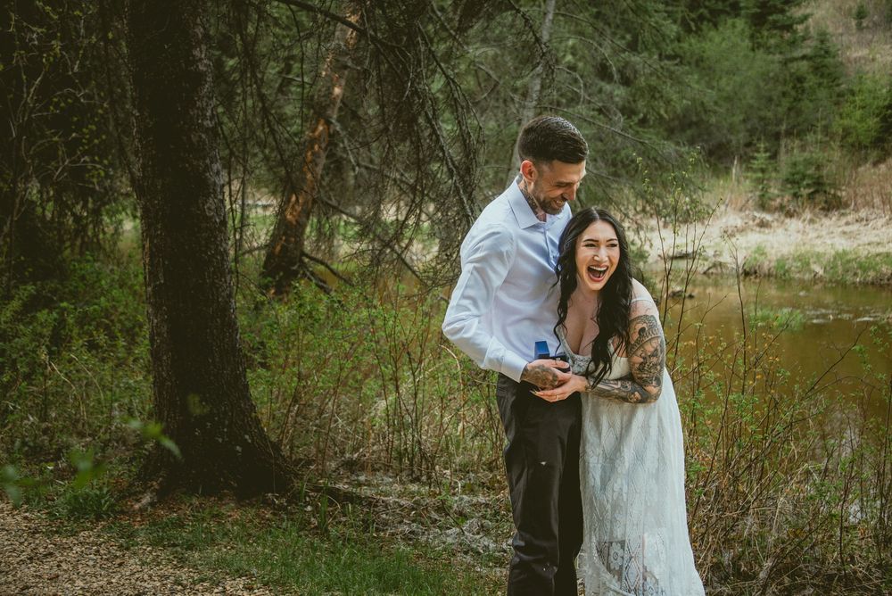 Couple laughing joyfully during outdoor proposal in forest setting near stream, man showing ring box to surprised woman.