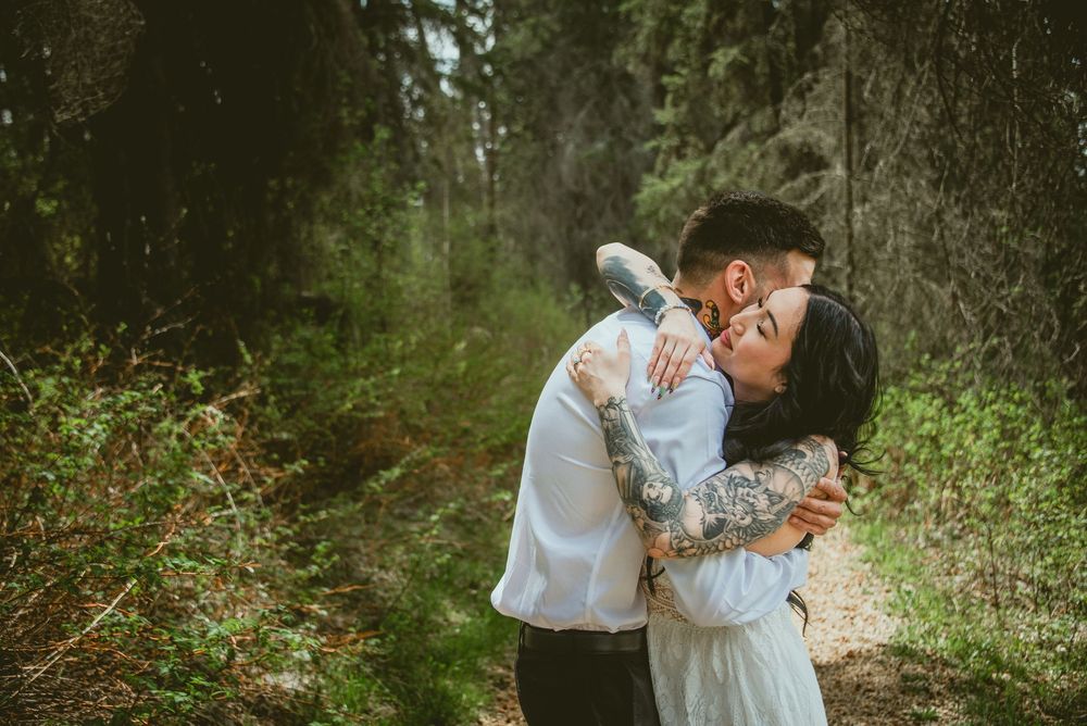 Couple shares romantic embrace on forest path, bride in white dress with tattoos hugging groom.