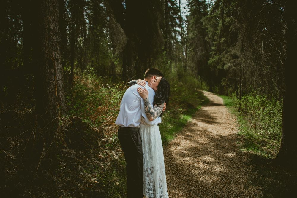 Couple sharing a romantic kiss on a forest path, bride in lace wedding dress surrounded by tall trees.