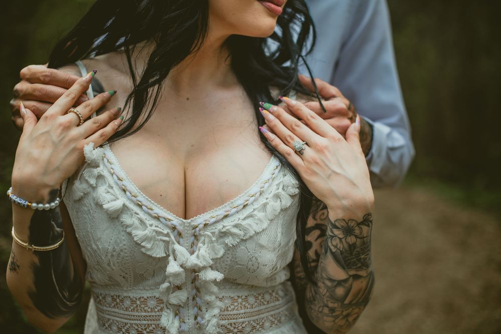 Close-up of bride in lace wedding dress with tattoos, wearing engagement ring, groom's hands on her shoulders.
