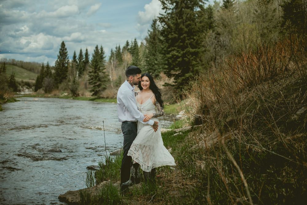 Couple embracing by a mountain river in a lush forest, bride in lace dress, groom in white shirt.