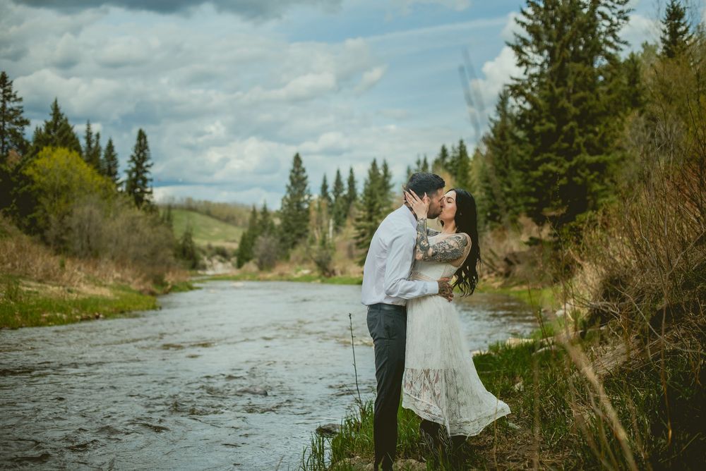 Couple kissing by a scenic mountain river surrounded by evergreen trees, bride in elegant lace gown.
