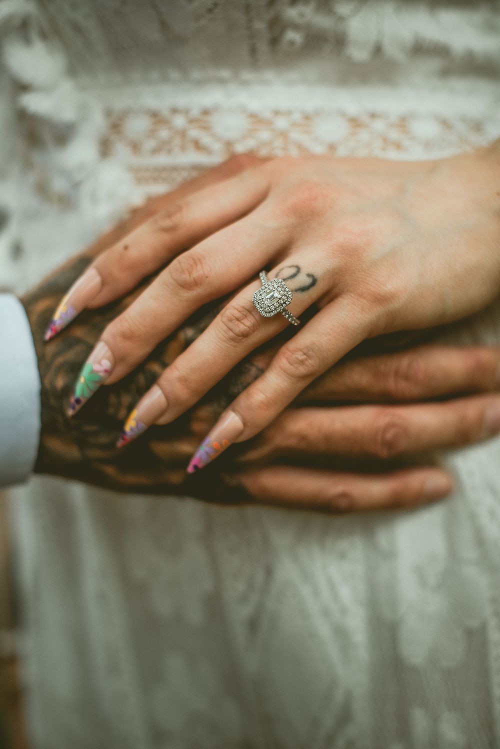 Close-up of a diamond engagement ring on a bride's hand with colorful nails, resting on a tattooed hand.