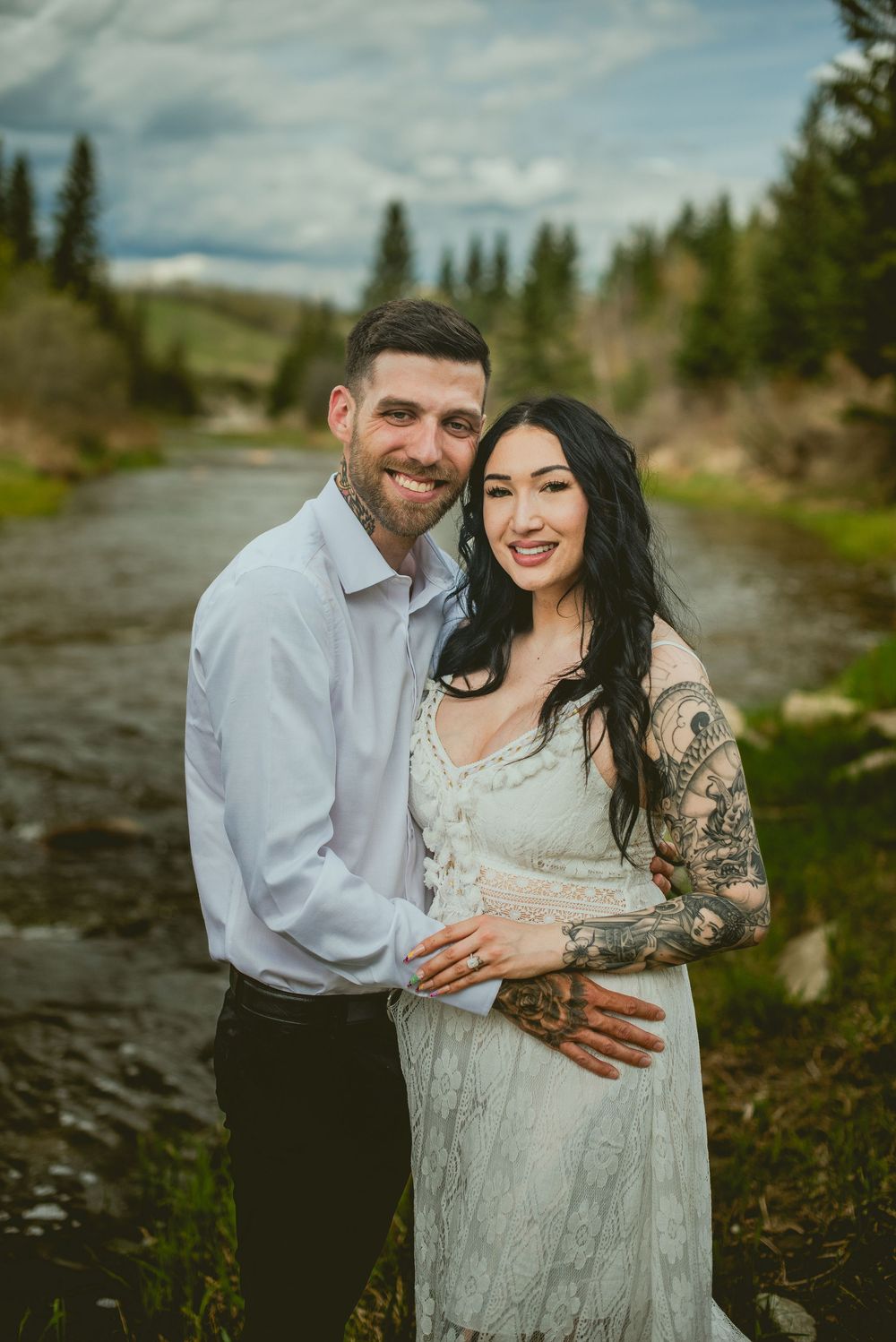Smiling couple poses by a mountain stream, woman in white lace dress with tattoos, man in light blue shirt.