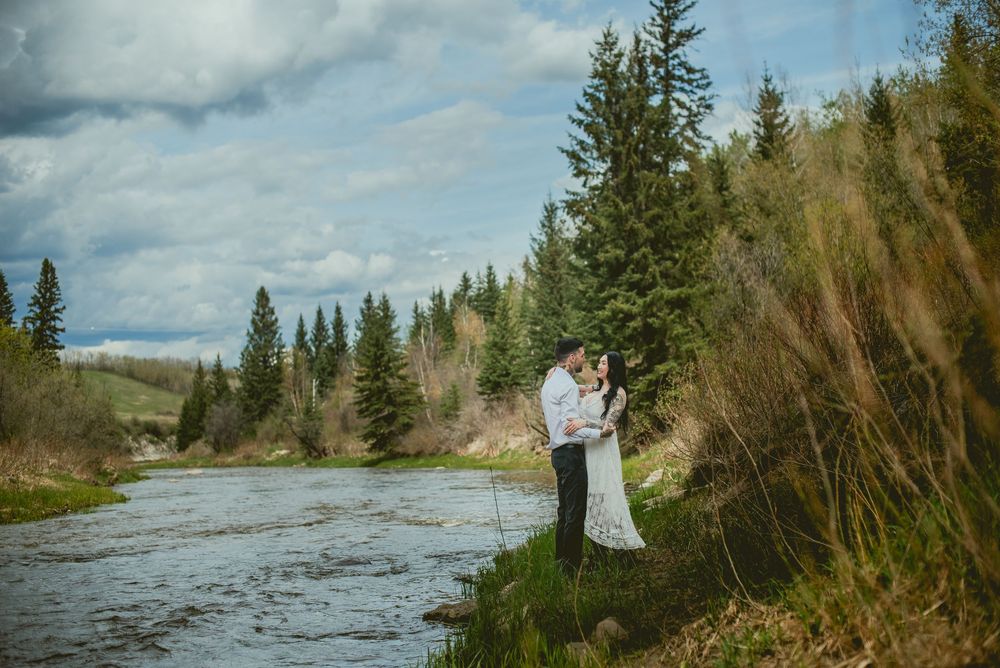 Bride and groom embrace by a scenic mountain river surrounded by evergreen trees under a cloudy sky.