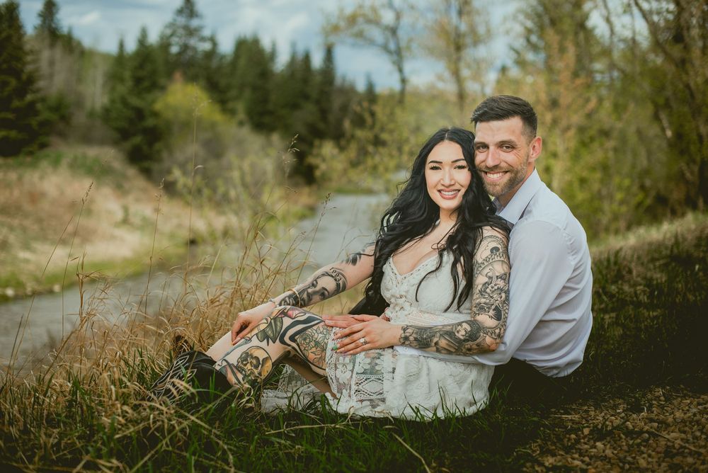 Tattooed couple sits together smiling in a lush outdoor setting beside a winding path.
