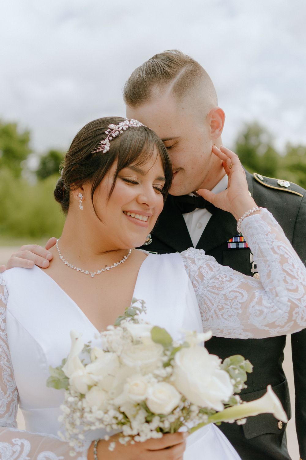 Military groom kisses bride's cheek as she smiles holding white rose bouquet in elegant lace wedding gown outdoors.