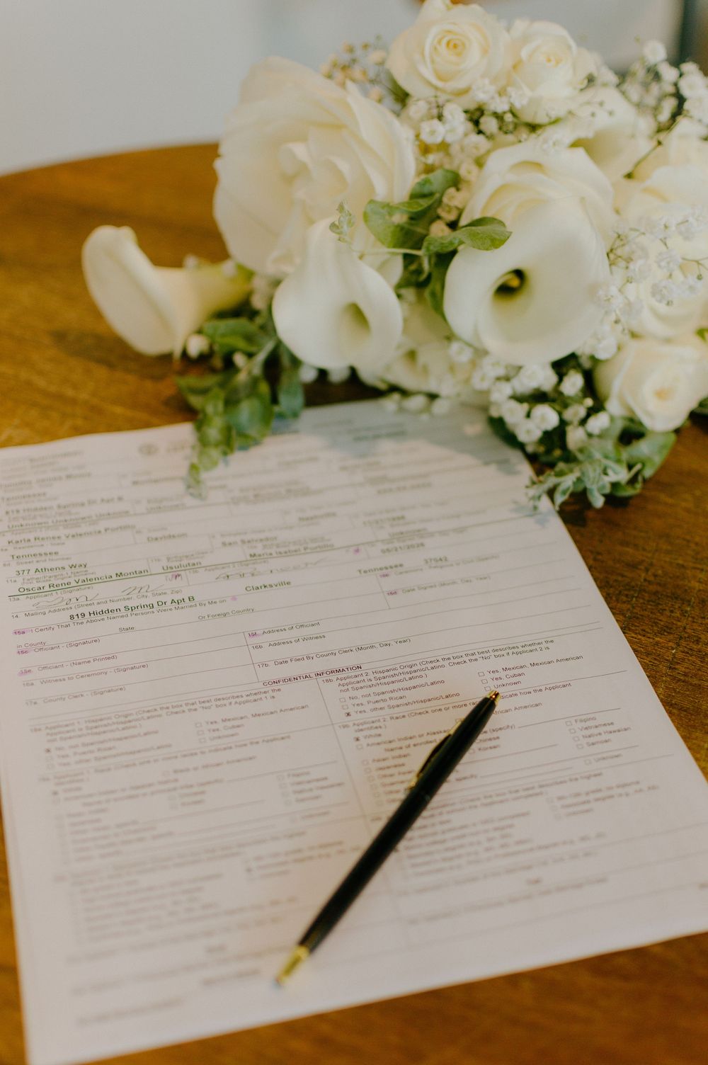 White wedding bouquet with roses and calla lilies beside a marriage license document and black pen on wooden table.