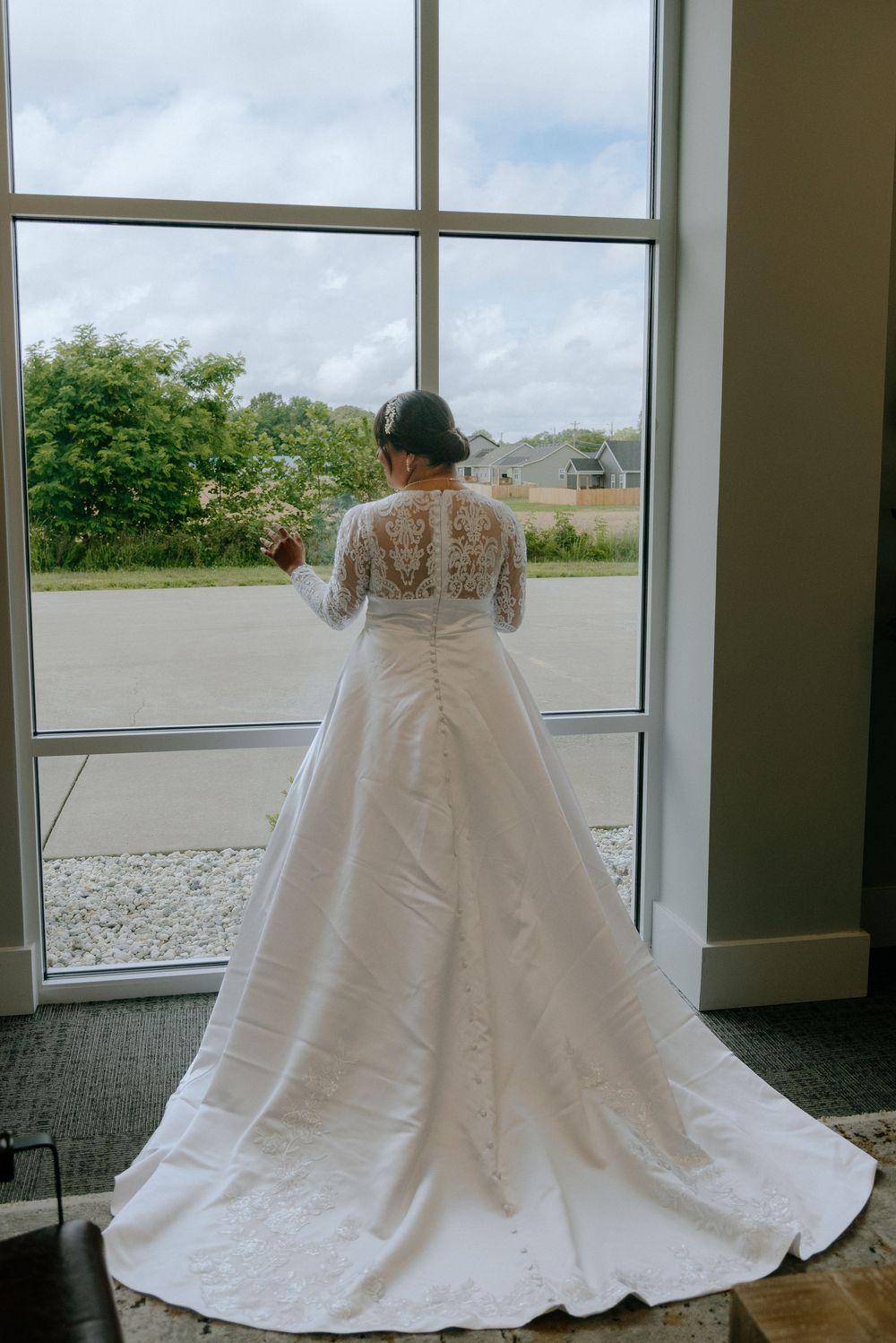 Bride in a white lace long-sleeve wedding gown with a train stands before a large window, holding flowers.